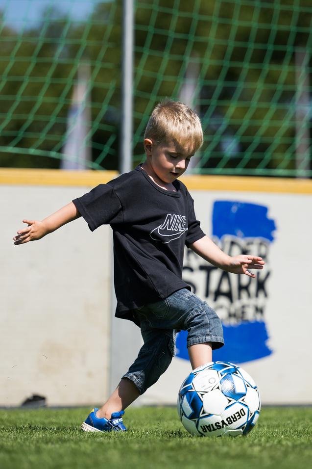 Kleiner Junge in schwarzem T-Shirt kickt einen Fußball auf einem grünen Rasenspielfeld. Im Hintergrund sind Wände sichtbar.
