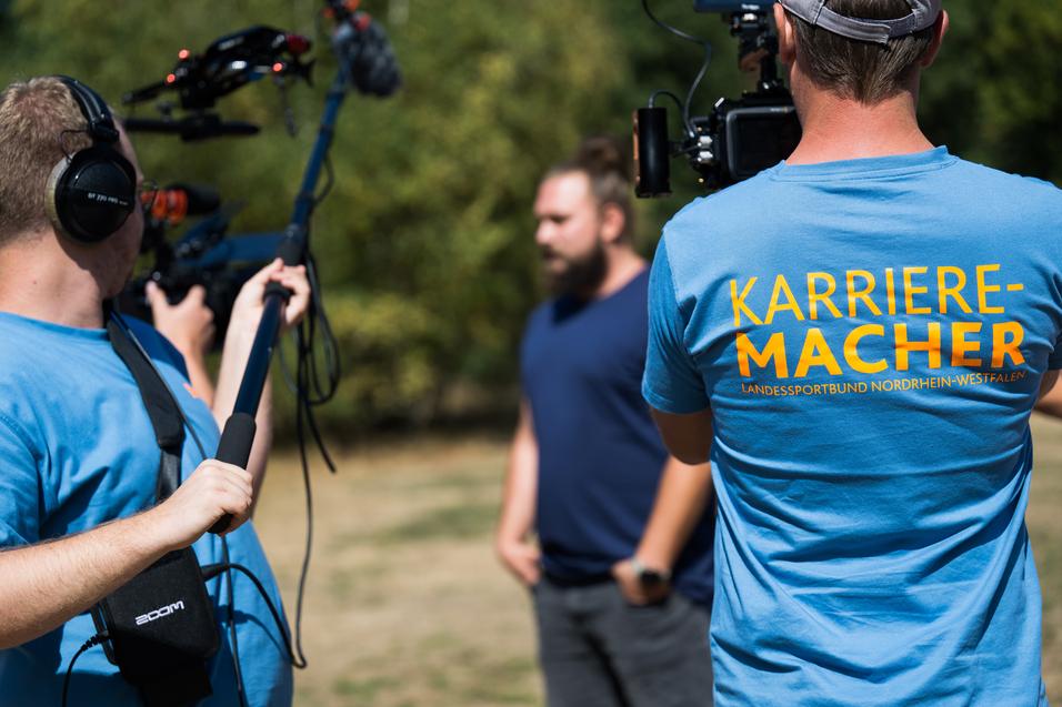 Drei Personen bei einer Videoaufnahme im Freien, einer trägt ein T-Shirt mit der Aufschrift „Karriere-Macher“.