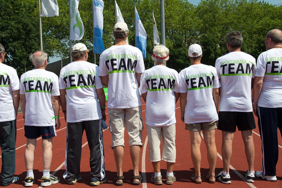 Gruppe von sieben Personen in weißen T-Shirts mit dem Aufdruck "TEAM" auf einer Laufbahn, Blick von hinten.