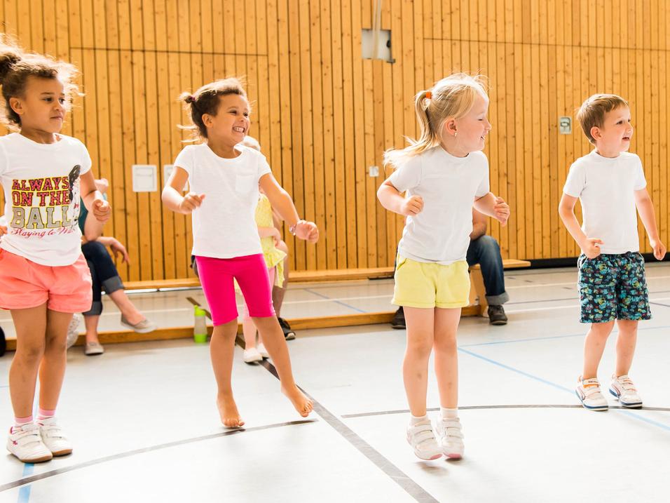 Fünf Kinder in T-Shirts springen fröhlich in einer Sporthalle mit Holzwänden und einen Zuschauer im Hintergrund.