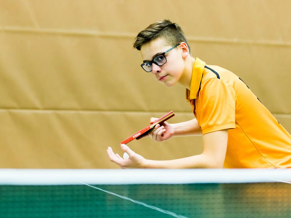 Ein junger Tischtennisspieler in orangem Shirt bereitet sich mit Schläger in der Hand auf den Aufschlag vor.