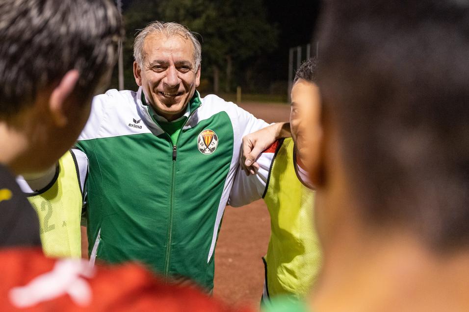 Trainer umringt von Spielern in sportlichen Trikots während einer Besprechung auf dem Fußballplatz bei Nacht.