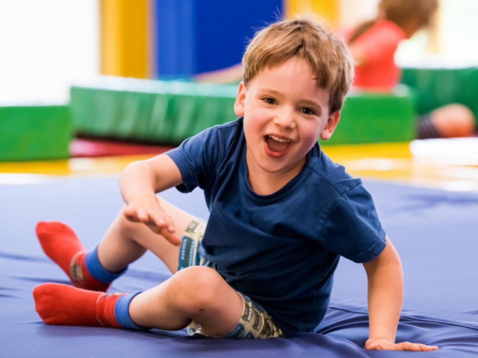 Ein fröhlicher Junge spielt auf einer blauen Matte in einer Freizeitstätte, umgeben von bunten Matten.