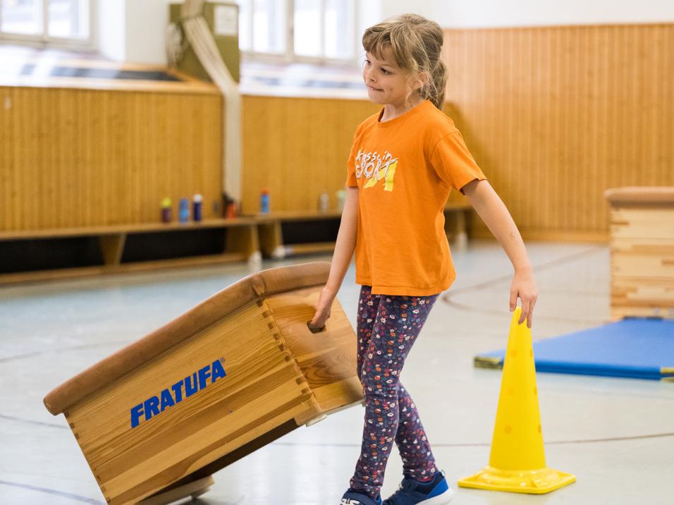 Mädchen in orangem T-Shirt trägt einen Turnkasten in einer Turnhalle, umgeben von gelben Hütchen und Matten.