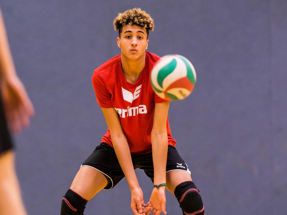 Junger Volleyballspieler in rotem Shirt bereit, einen bunten Volleyball zu empfangen, auf einem Holzsportboden.