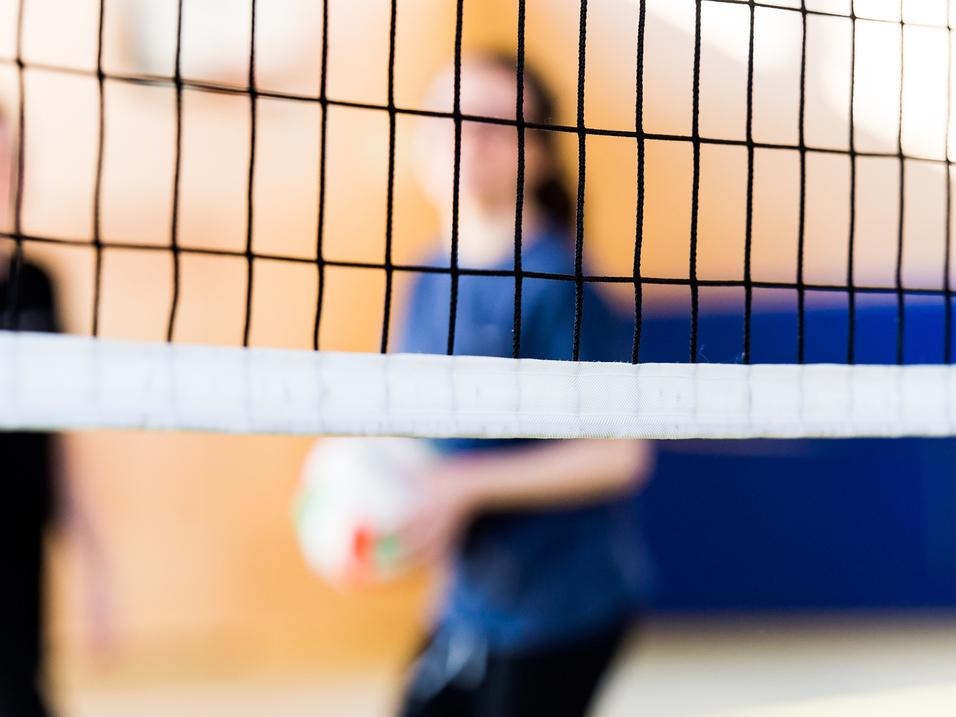Unscharfe Silhouetten von zwei Personen hinter einem Volleyballnetz in einer Sporthalle. Eine Person hält einen Volleyball.